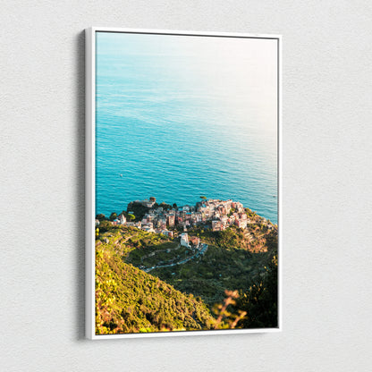 Framed canvas wall art featuring sunlit coastal town with green hills and calm blue ocean background.