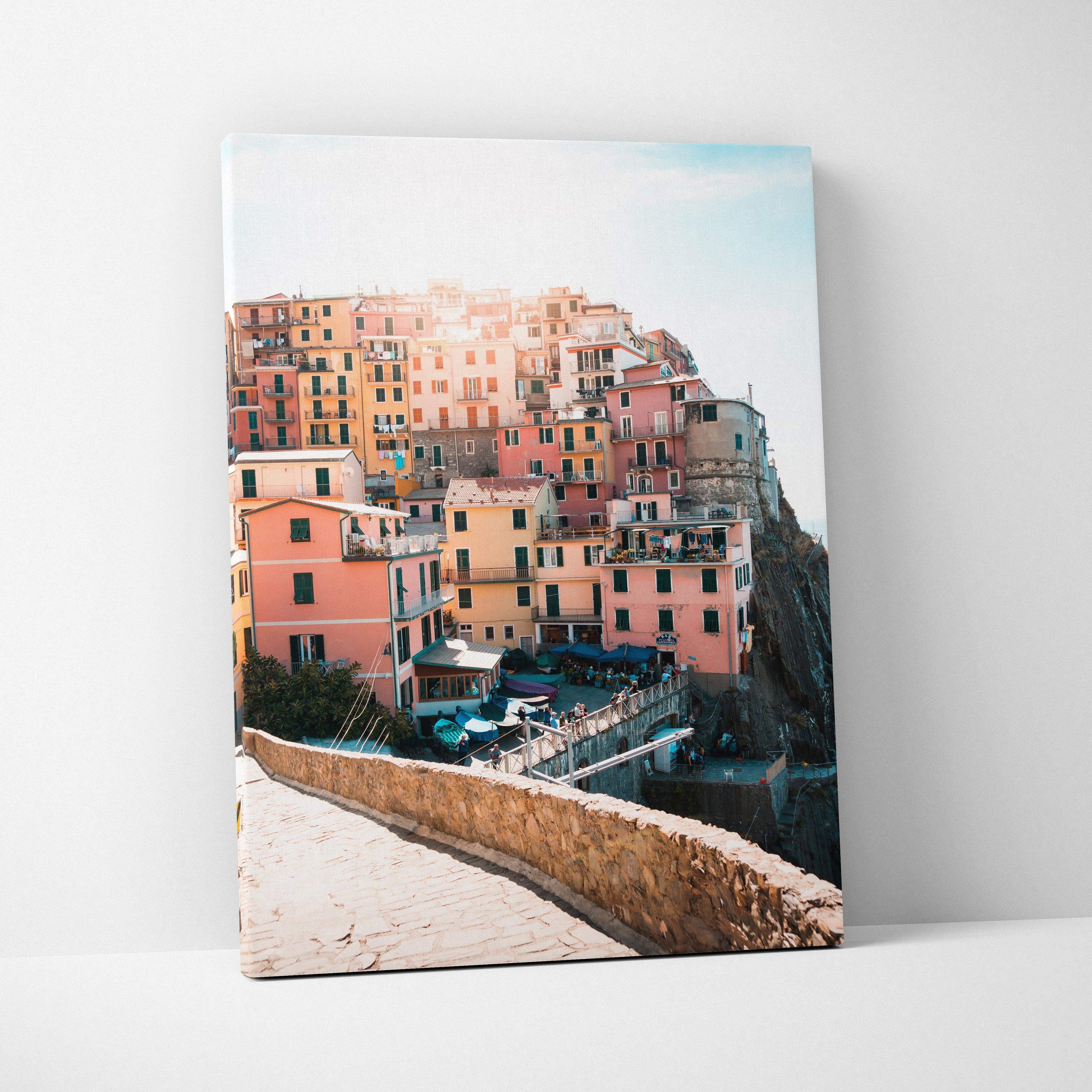 Canvas wall art featuring a colorful coastal village with stacked pastel buildings and a stone walkway in soft sunlight.