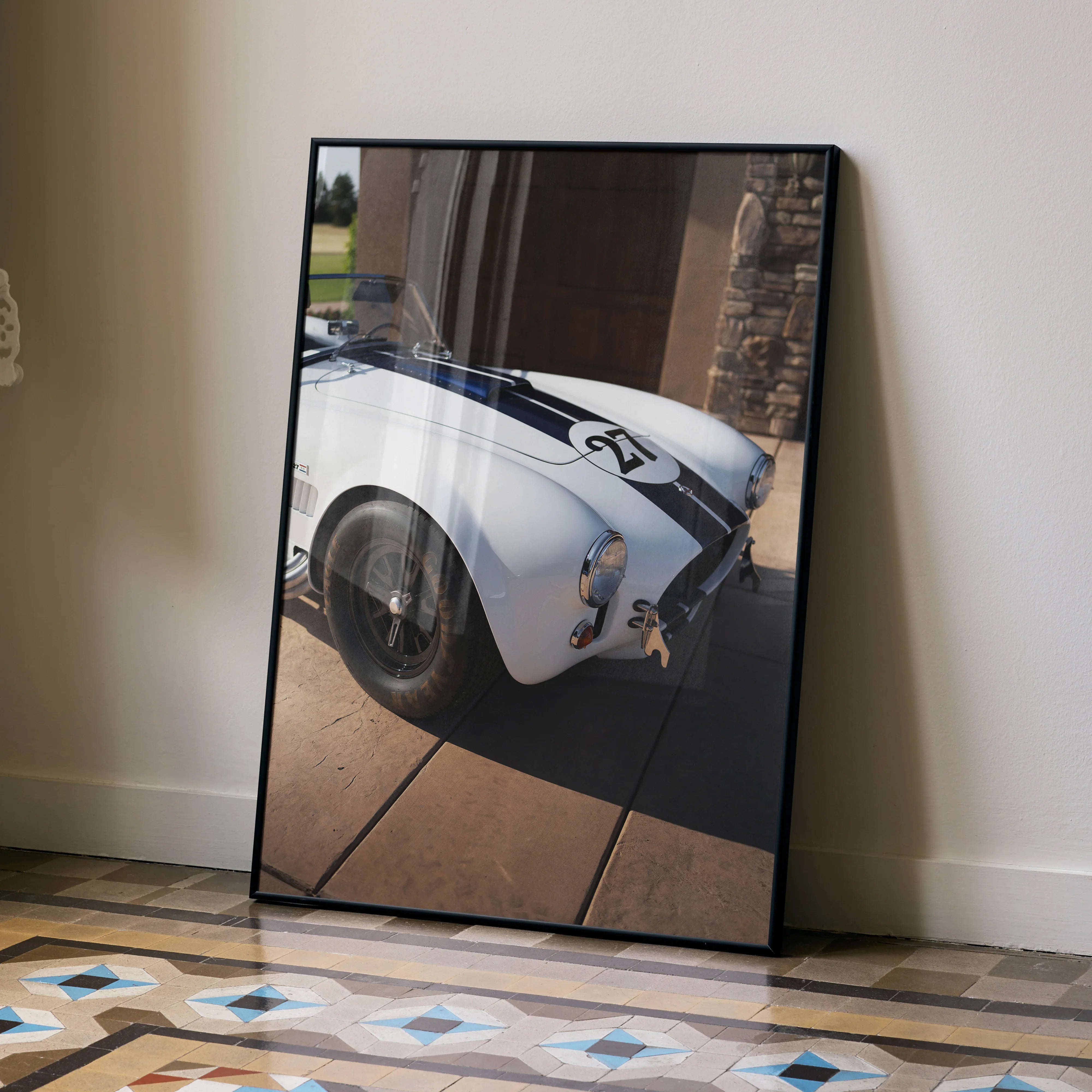 Shelby Cobra car poster with white body and black racing stripes leaning against beige wall wall art.
