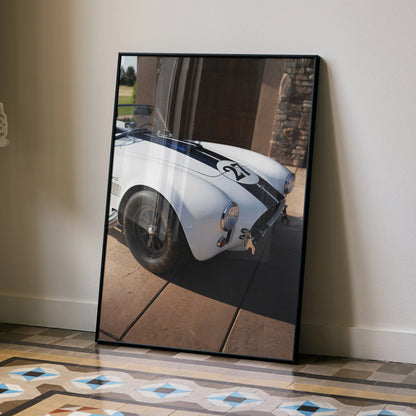 Shelby Cobra car poster with white body and black racing stripes leaning against beige wall wall art.