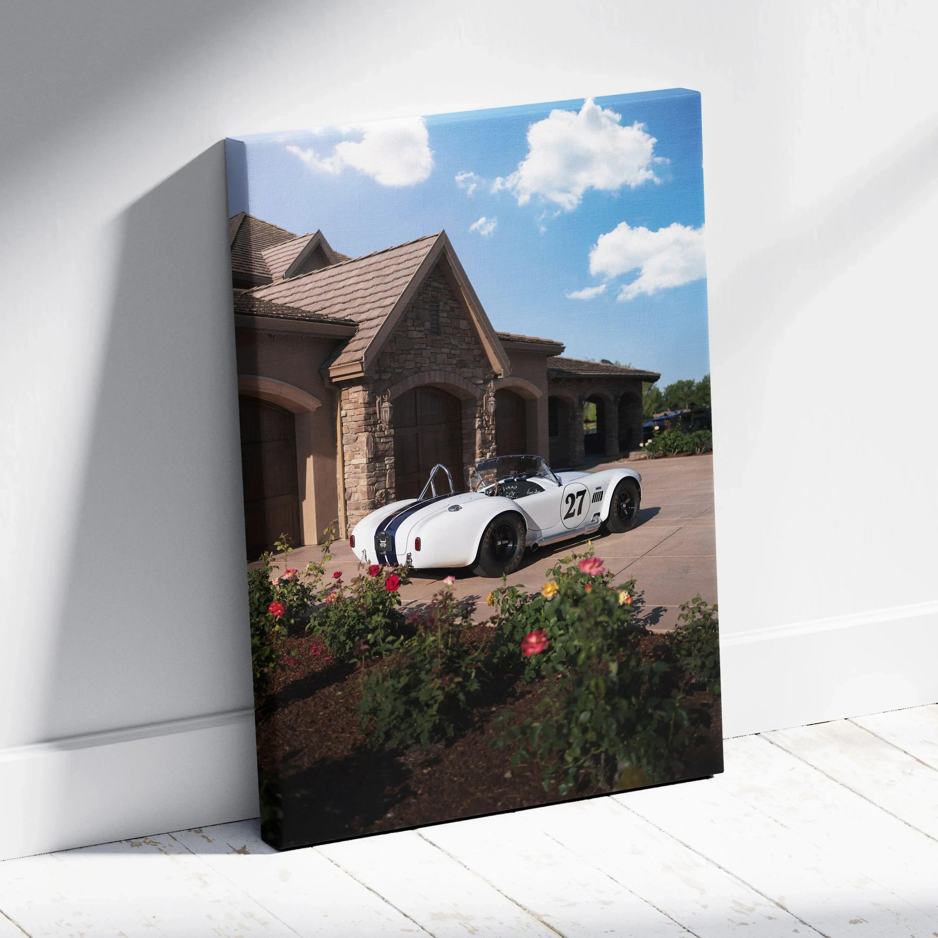 Vertical canvas wall art featuring a white Shelby Cobra parked in front of a stone house under a bright blue sky.