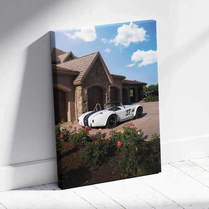 Vertical canvas wall art featuring a white Shelby Cobra parked in front of a stone house under a bright blue sky.