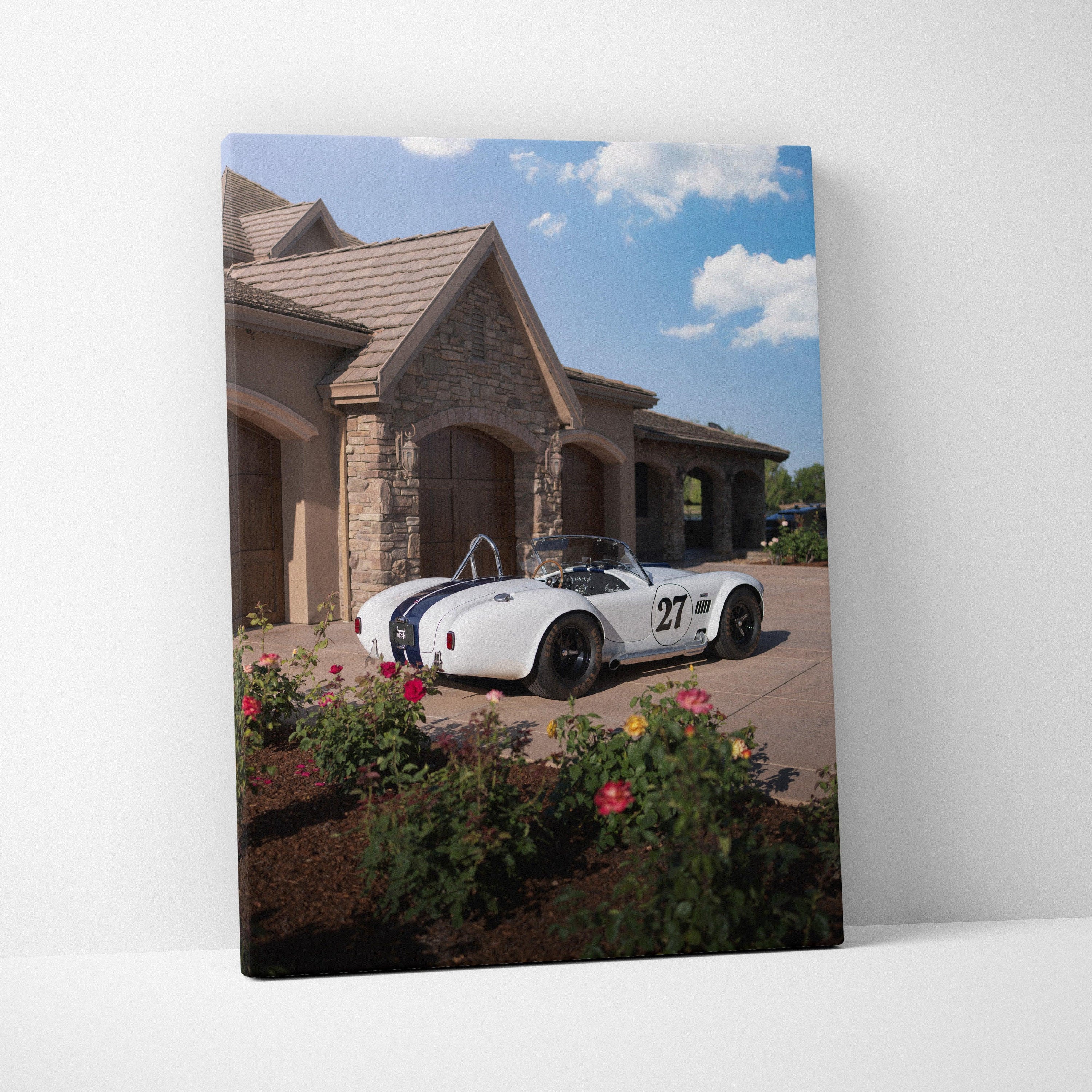 Vertical canvas wall art featuring a white Shelby Cobra sports car parked by a stone house under a blue sky.
