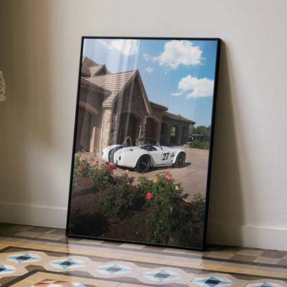 Shelby Cobra car poster leaning against wall with blue sky and brick house background, wall art.