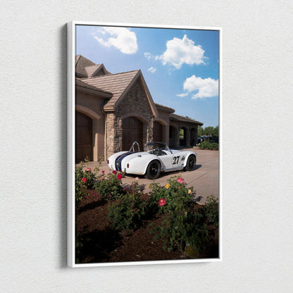 Vertical canvas art of white Shelby Cobra sports car parked by stone house under blue sky with clouds.