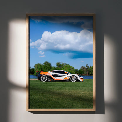 McLaren 570S poster wall art featuring a white and orange sports car on green grass under a bright blue sky with clouds.