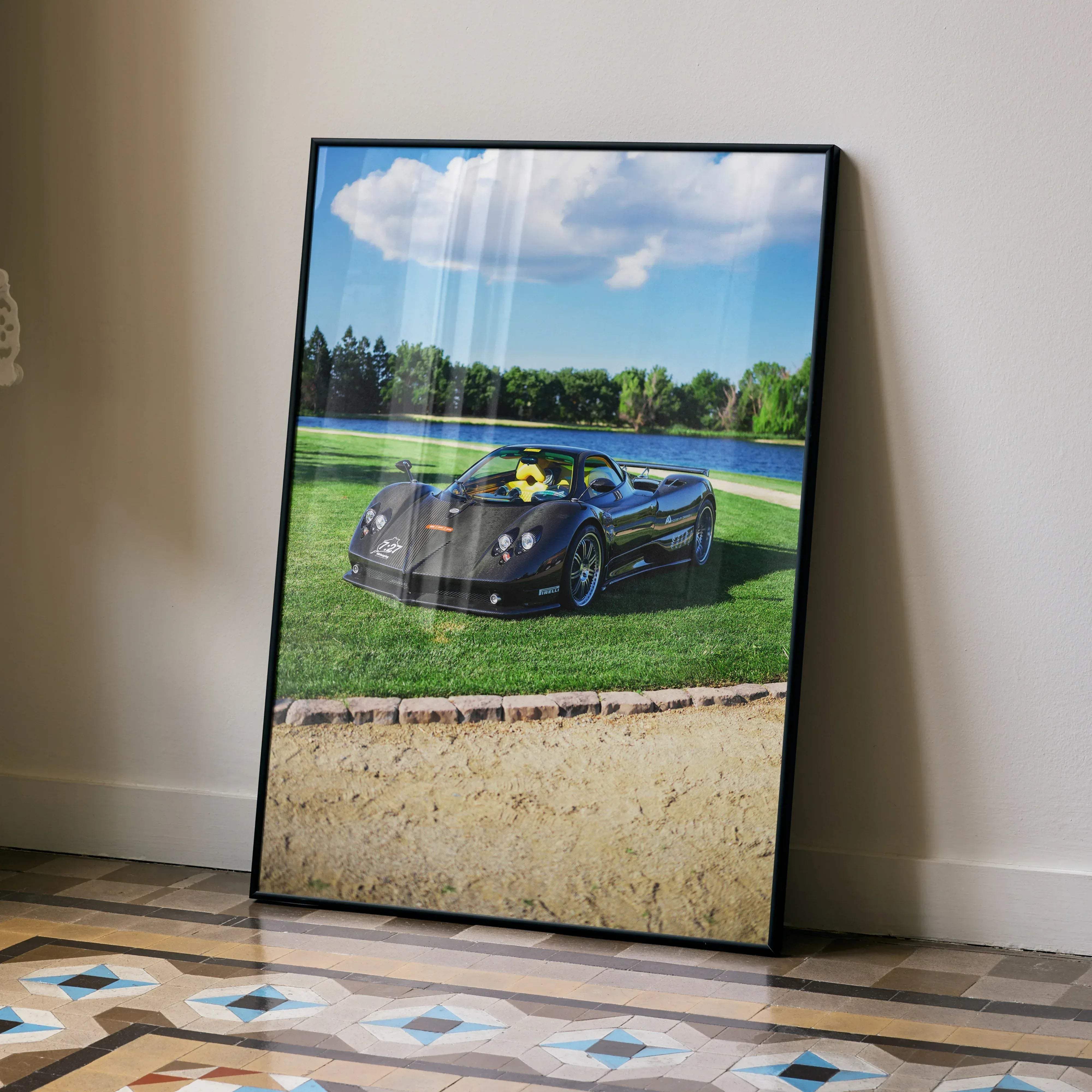 Black framed poster of a glossy black Pagani Zonda F Clubsport with yellow interior parked on grass by water under blue sky
