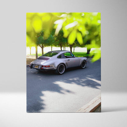 Vertical canvas art of a classic silver Porsche 911 parked on a sunlit street with green foliage overhead.