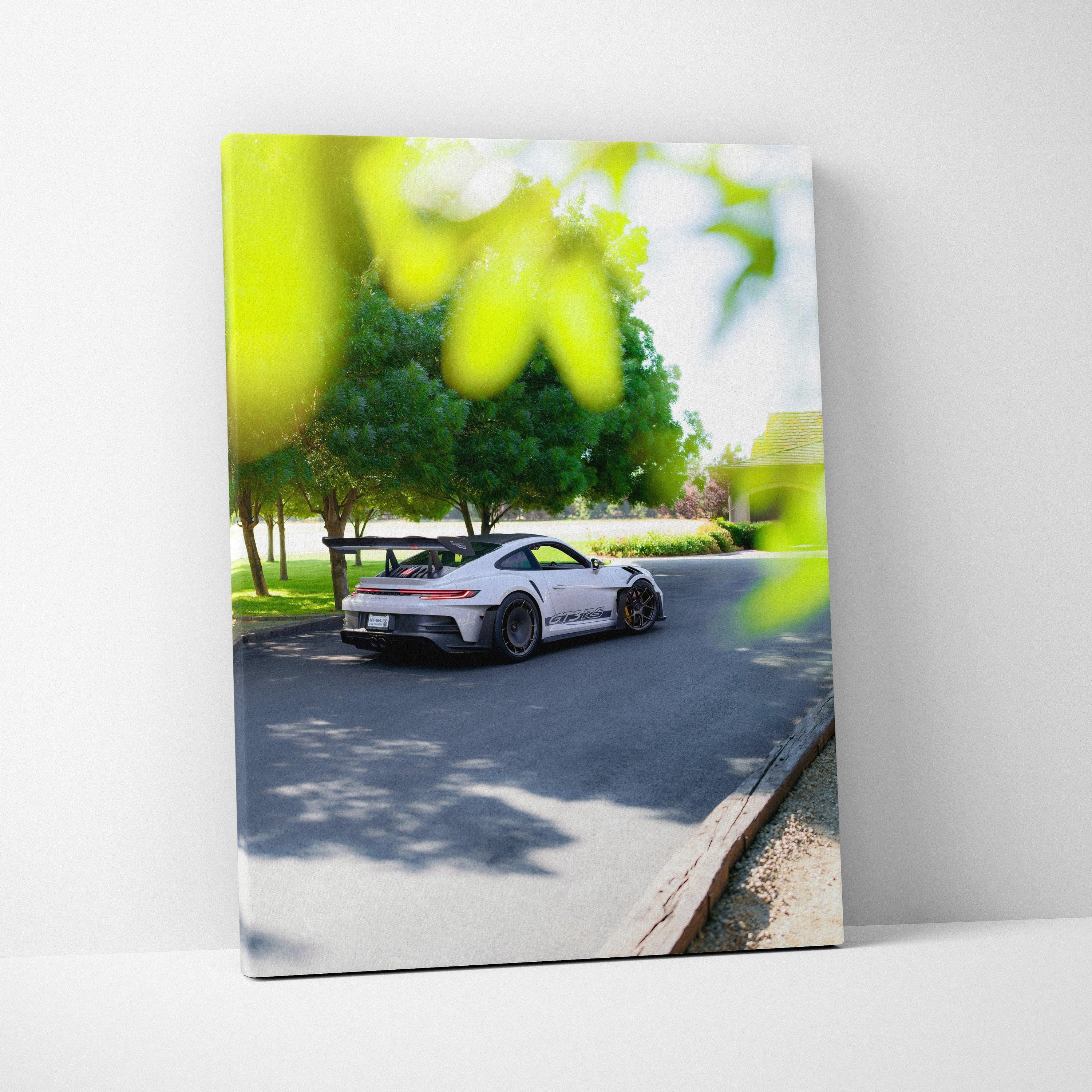 Vertical canvas art of white Porsche 911 GT3 RS parked on sunlit street with green trees background.