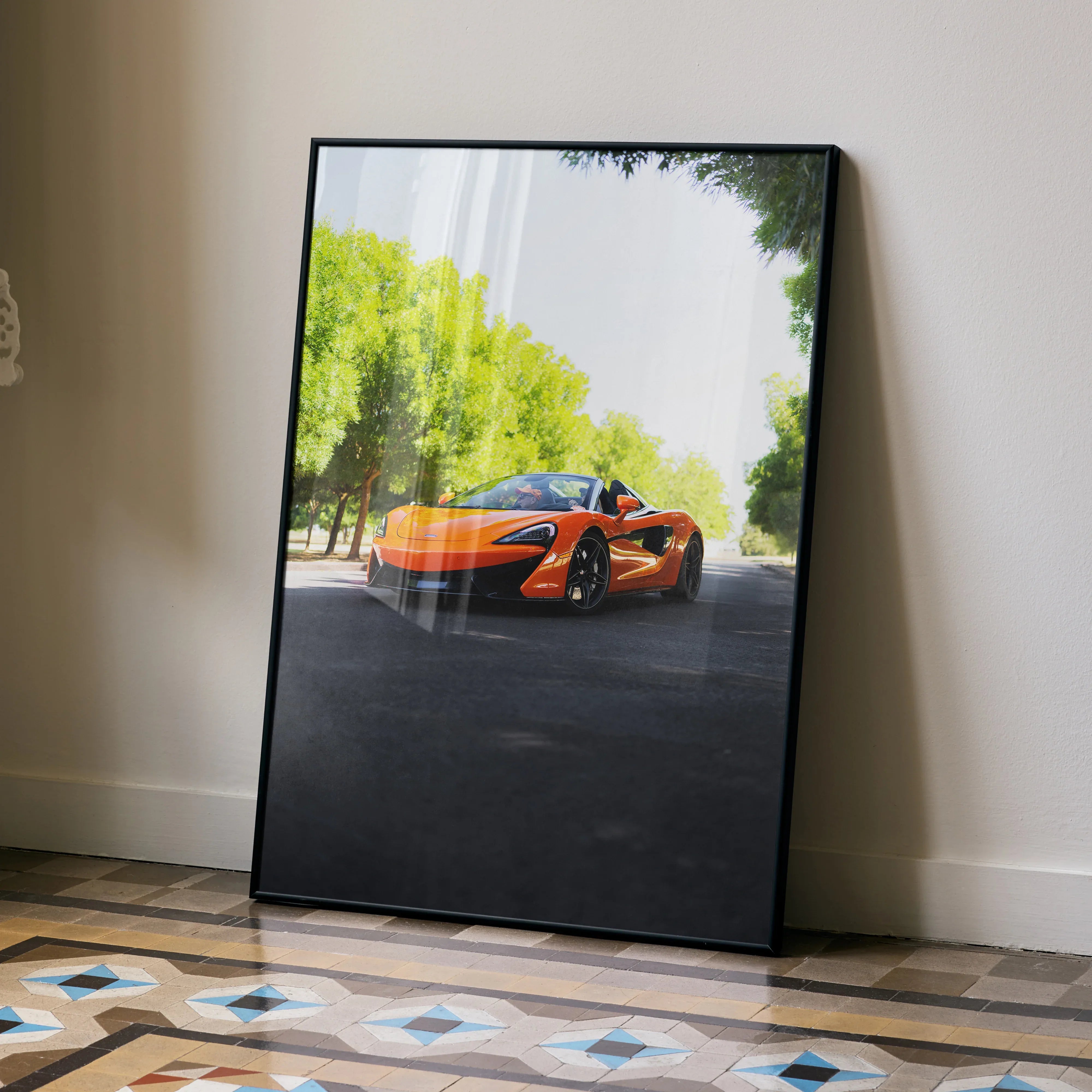 McLaren 570S poster wall art featuring bright orange sports car on sunlit road with green trees.