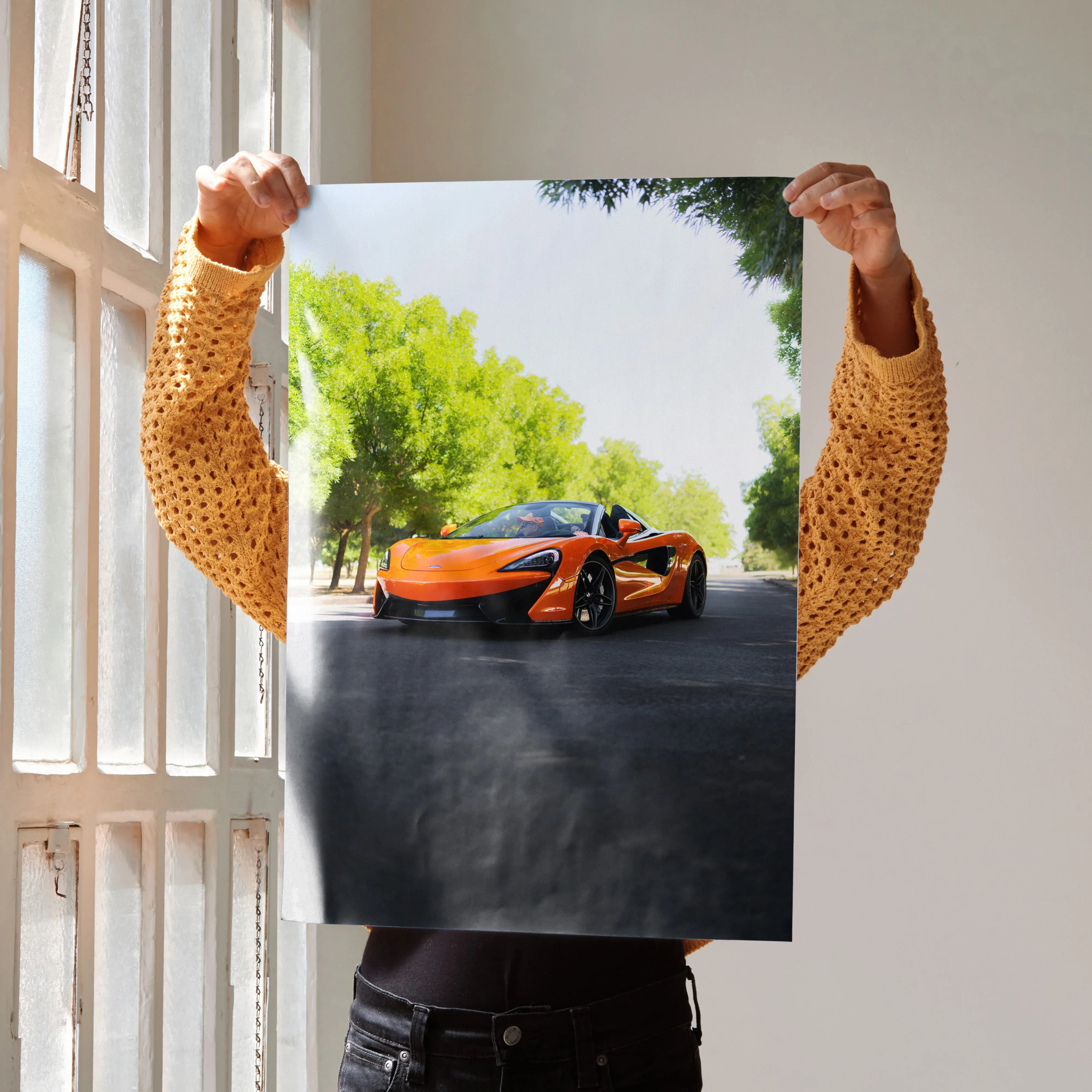 McLaren 570S orange sports car poster wall art held by person near window.