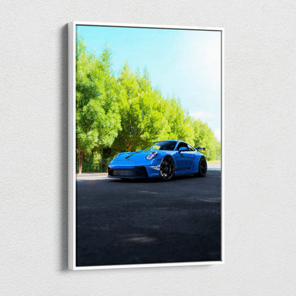 Vertical canvas wall art featuring a blue Porsche 911 GT3 parked on sunlit asphalt beside green trees.