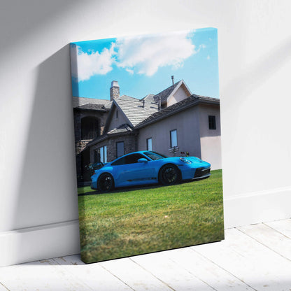 Vertical canvas wall art featuring a blue Porsche 911 GT3 parked on grass in front of a house under a blue sky.