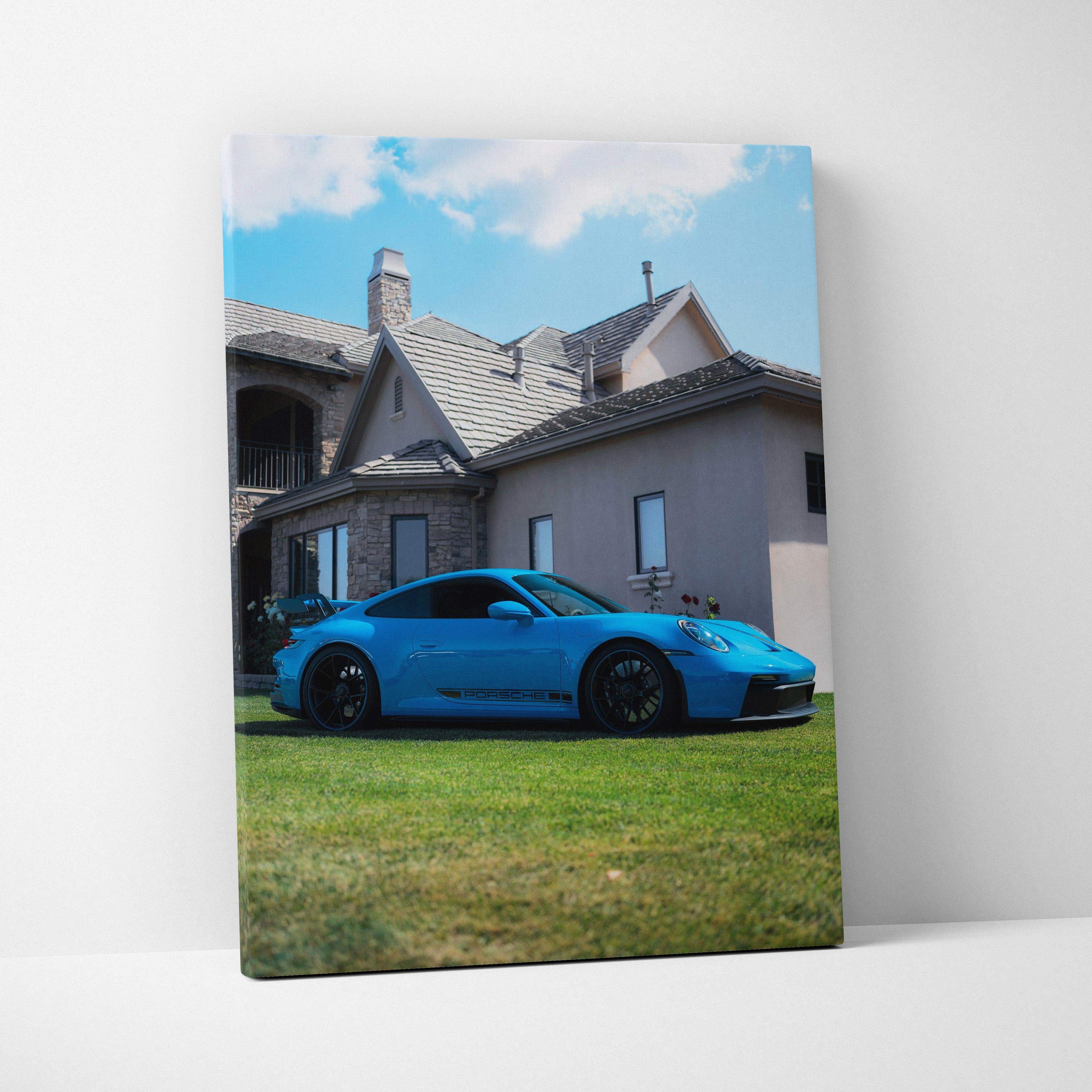 Vertical canvas wall art featuring a blue Porsche 911 GT3 parked on grass in front of a house under a blue sky.