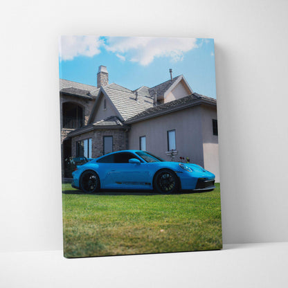 Vertical canvas wall art featuring a blue Porsche 911 GT3 parked on grass in front of a house under a blue sky.