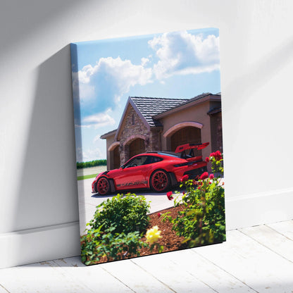 Vertical canvas wall art featuring a red Porsche 911 GT3 RS parked by a rustic house under a blue sky with clouds.