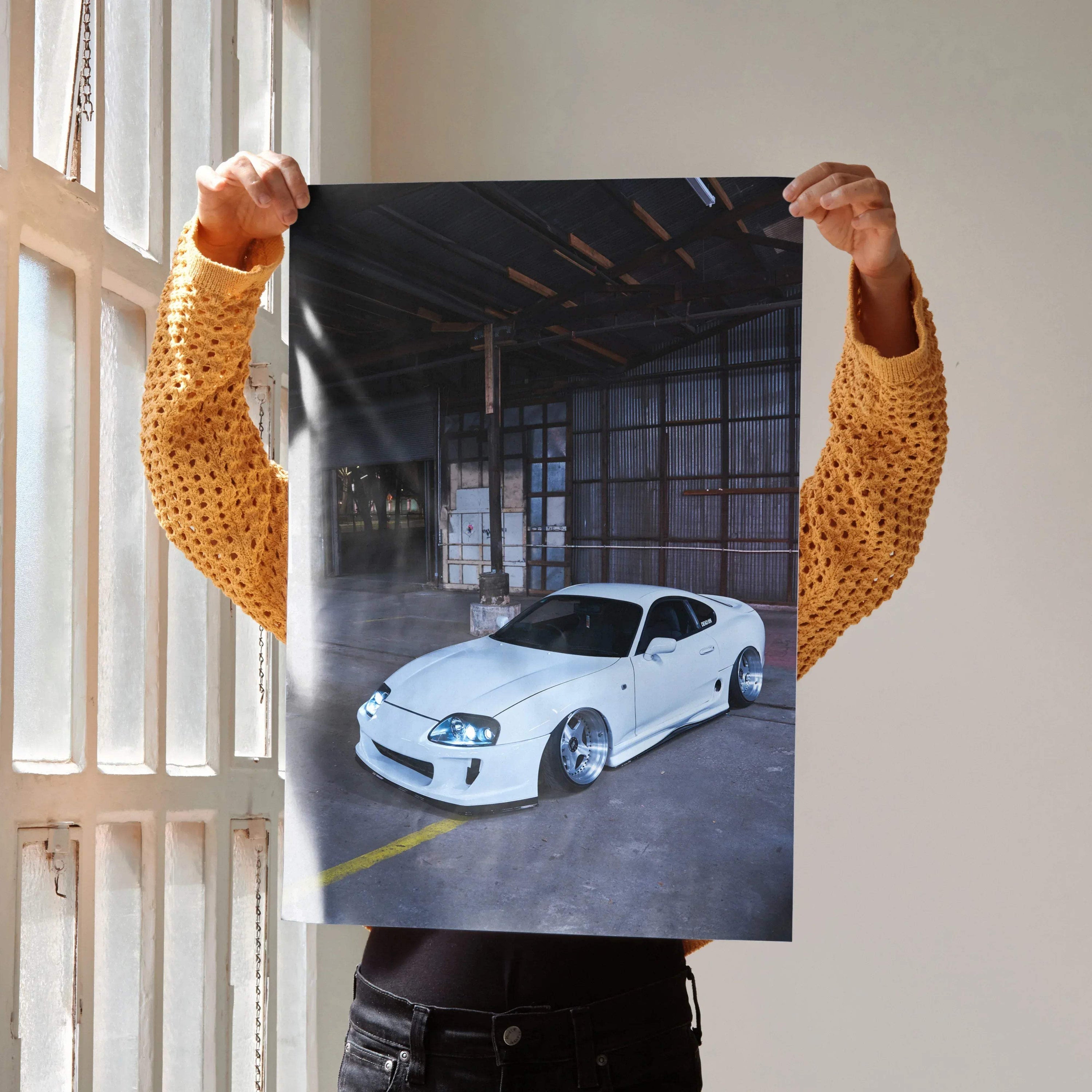 Toyota Supra MK4 white sports car poster held indoors, automotive wall art with warehouse background.