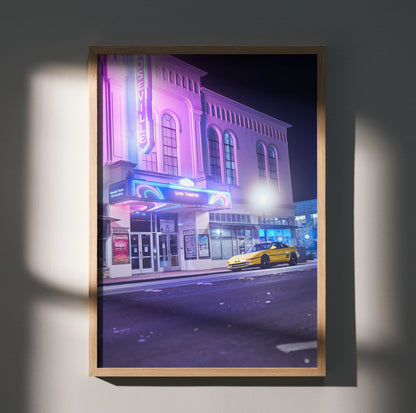 Toyota MR2 yellow sports car parked by neon-lit theater poster wall art in nighttime city.