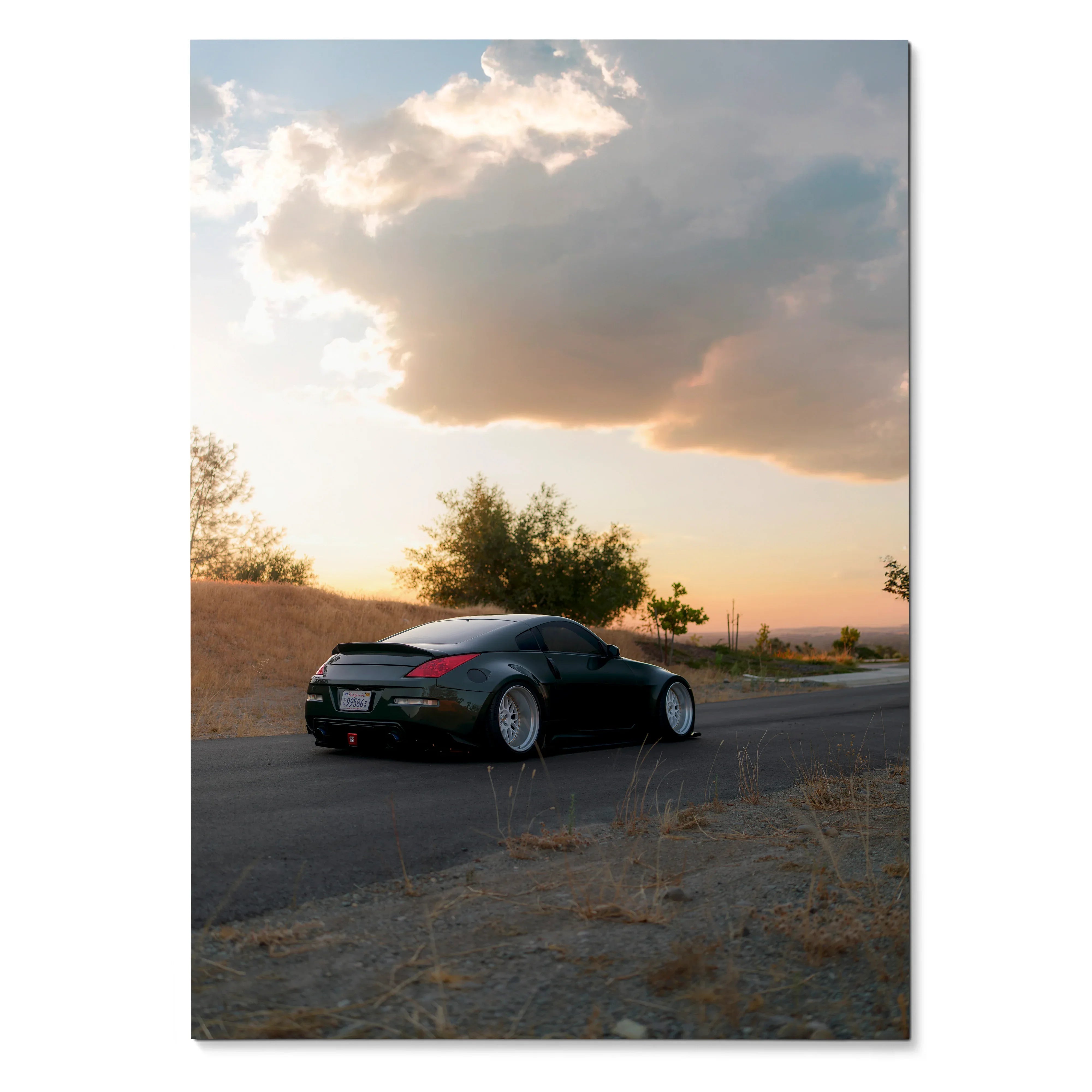 Nissan 350z black sports car on road at sunset poster wall art with cloudy sky background.