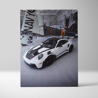 Vertical canvas art featuring a white Porsche 911 GT3RS with black racing stripes in a hangar wall art display.
