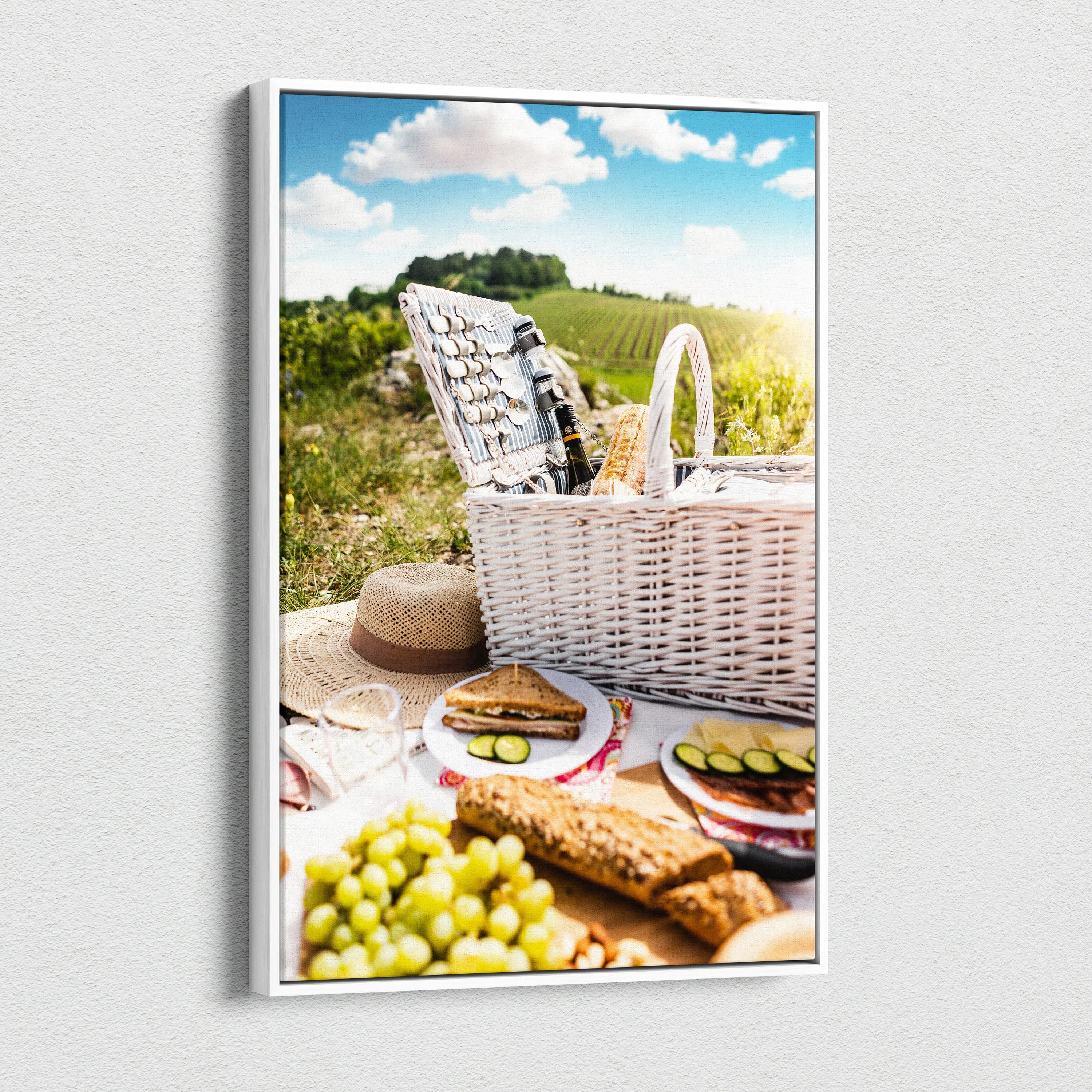 Vertical canvas wall art showing a picnic scene with white wicker basket, grapes, bread, and countryside decor.
