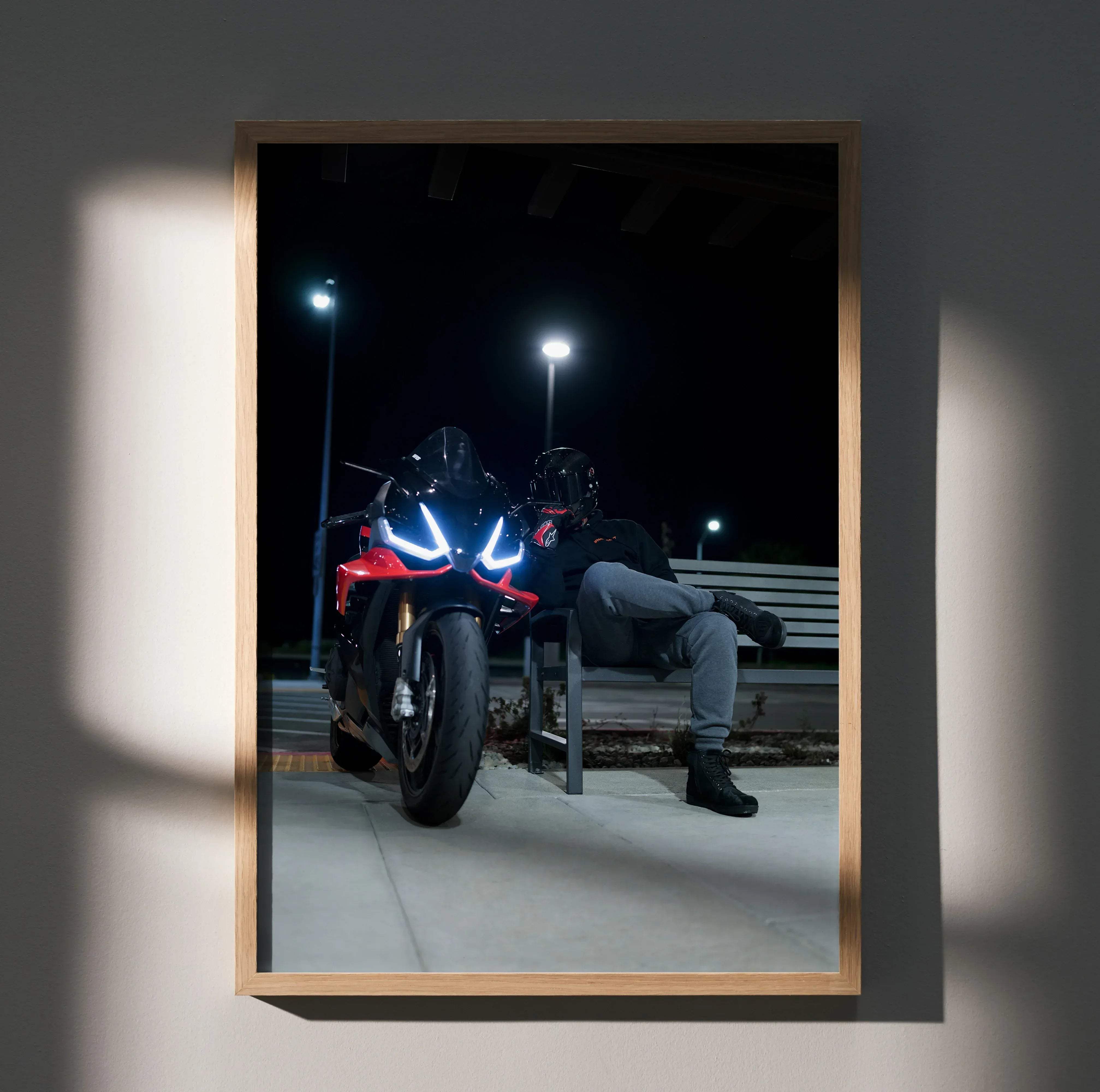 A framed photograph of a red and black aprilia rsv4 sport motorcycle with glowing headlights and a male wearing a shark aeron gp helmet sitting nearby