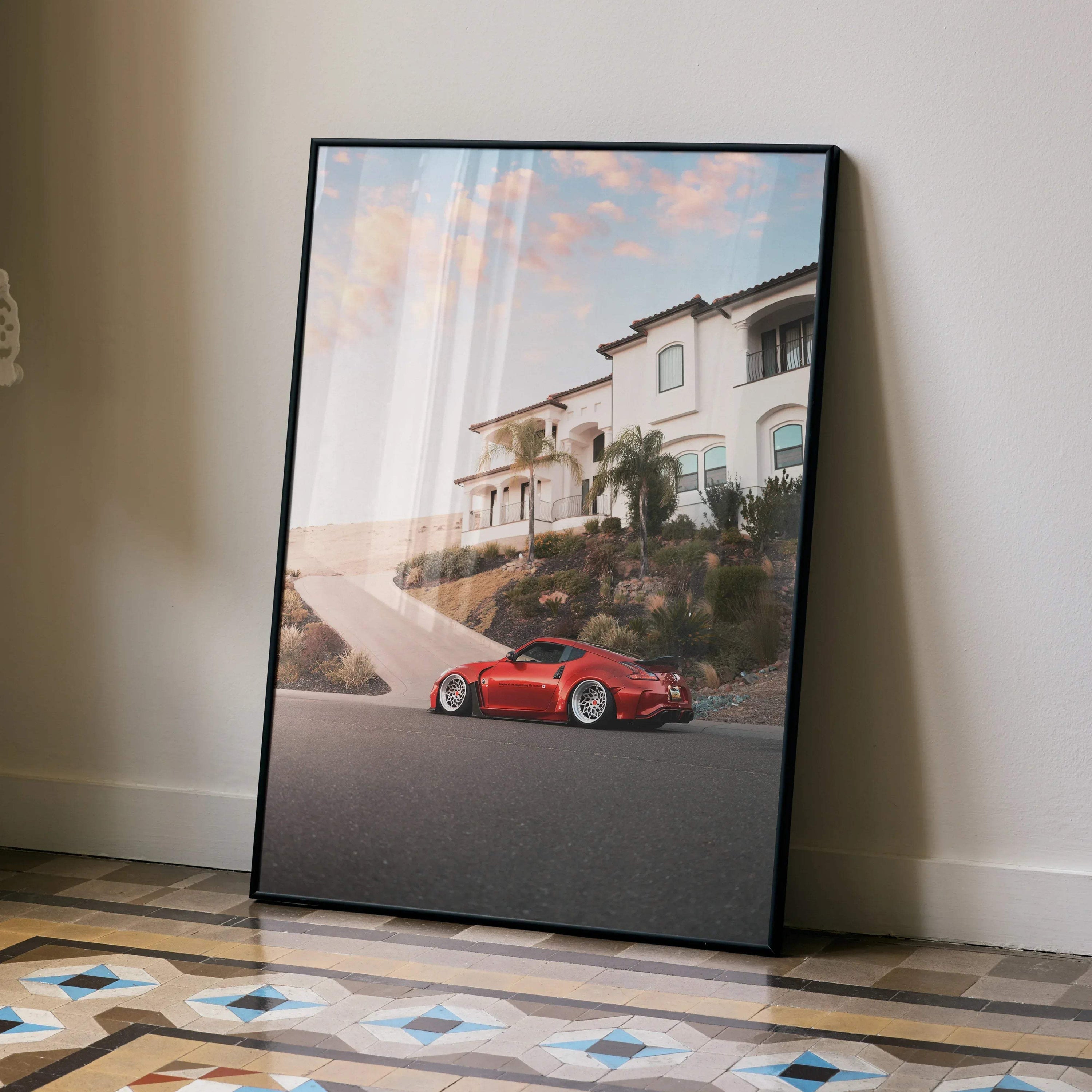 Red Nissan 370z parked by a white house at sunset poster wall art in black frame.