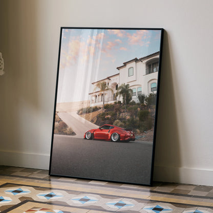 Red Nissan 370z parked by a white house at sunset poster wall art in black frame.