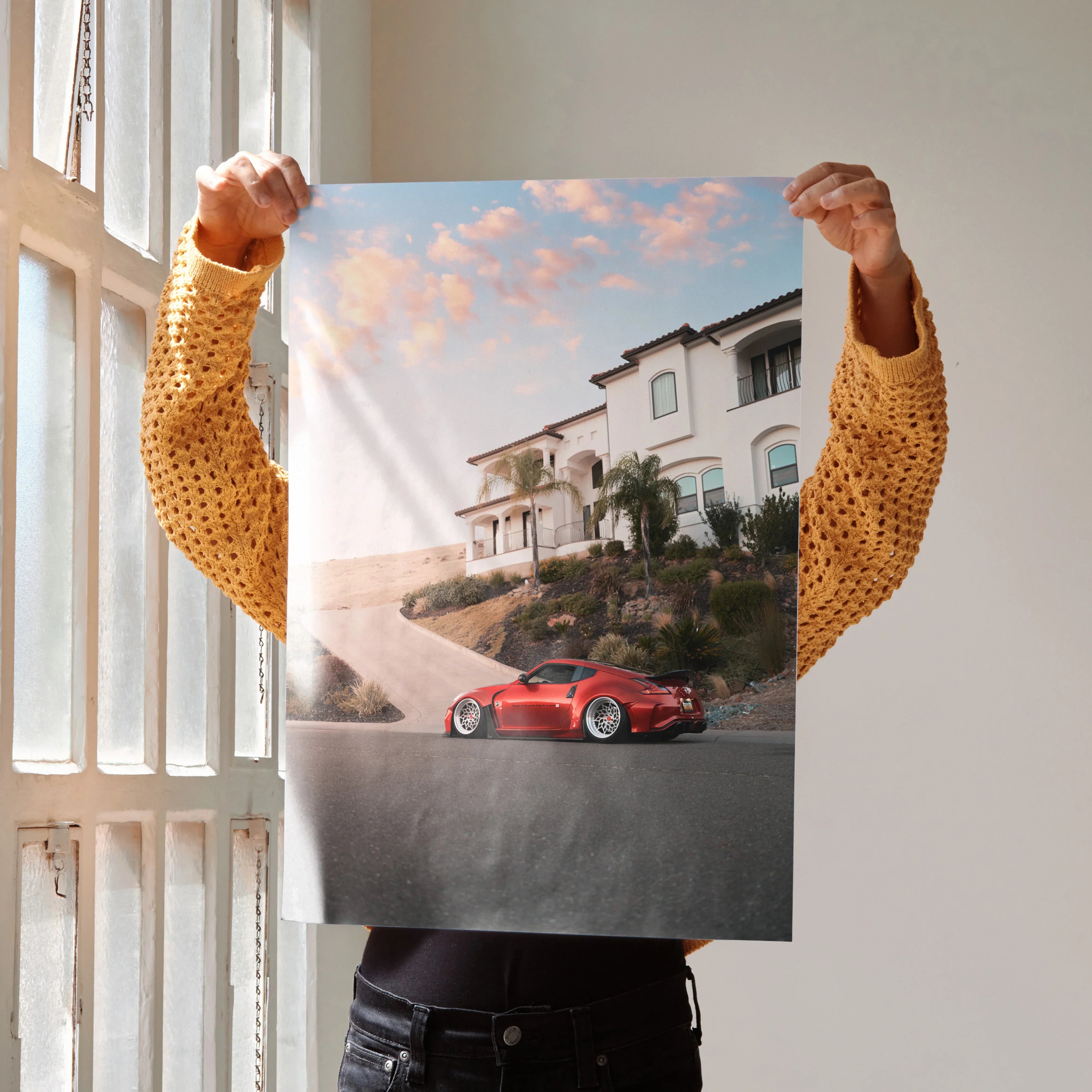 Nissan 370z poster wall art featuring a red car parked in front of a modern house under a sunset sky.