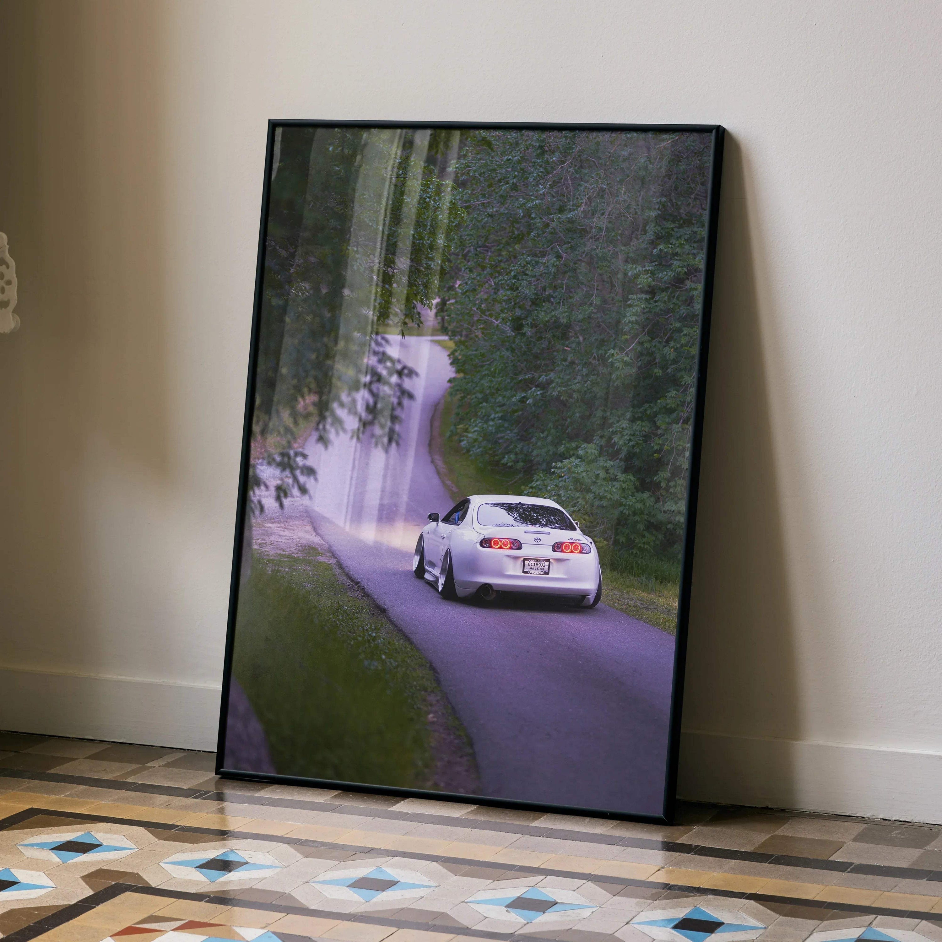 Toyota Supra MK4 driving on forest road poster wall art in black frame, leaning on wall.