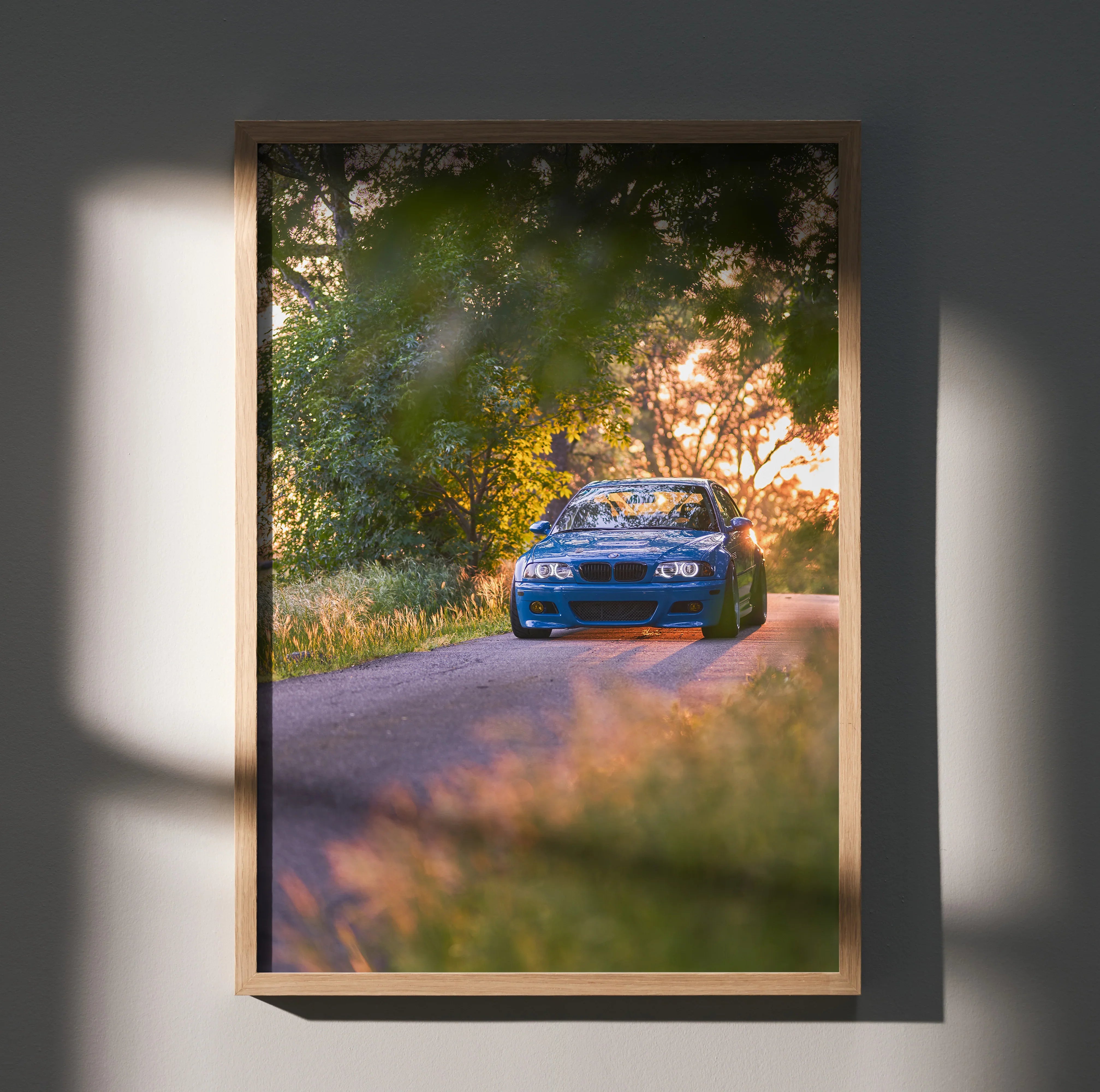 BMW E46 M3 driving on tree-lined road at sunset, vibrant poster wall art in wooden frame.