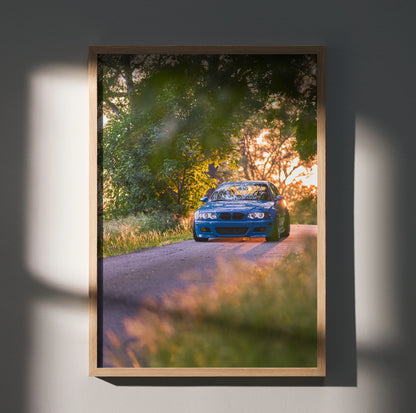 BMW E46 M3 driving on tree-lined road at sunset, vibrant poster wall art in wooden frame.