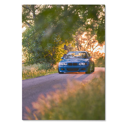 BMW E46 M3 driving on tree-lined road at sunset poster wall art with vibrant natural colors.