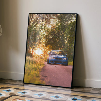 BMW E46 M3 driving on tree-lined road at sunset poster wall art in black frame.