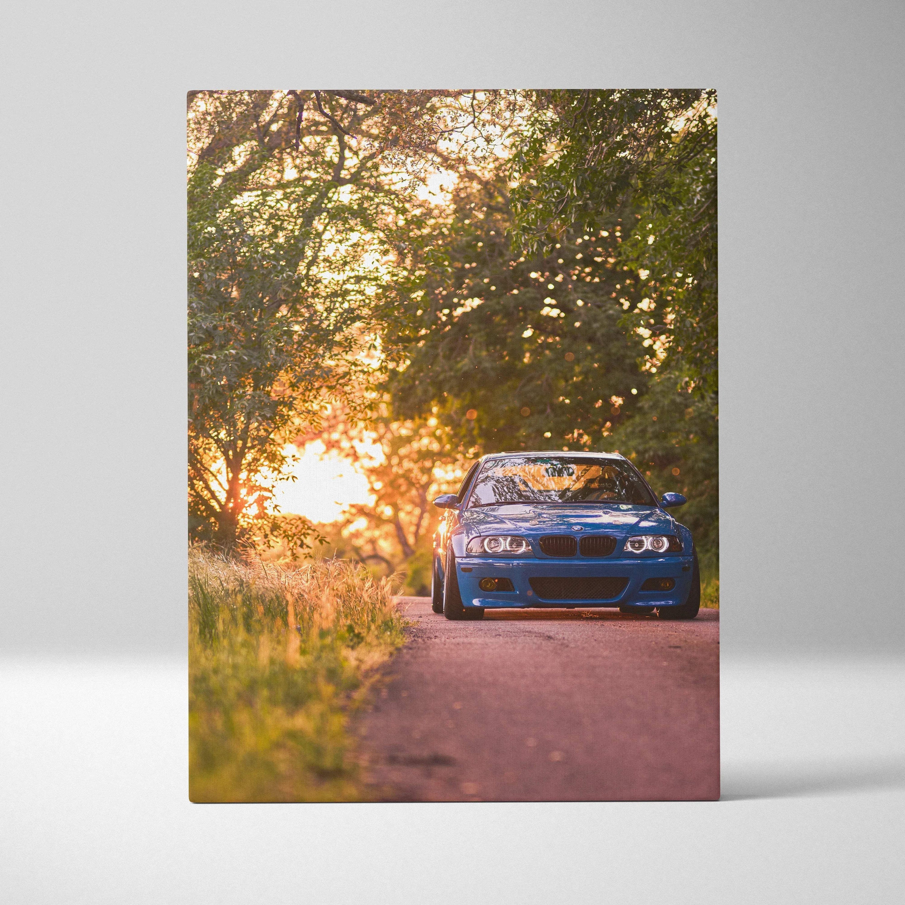 Vertical canvas wall art featuring a blue BMW E46 M3 on a sunlit forest road at sunset.