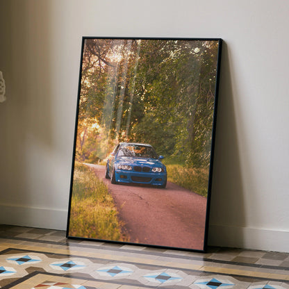 BMW E46 M3 poster with blue car driving on sunlit forest road, automotive wall art in black frame.