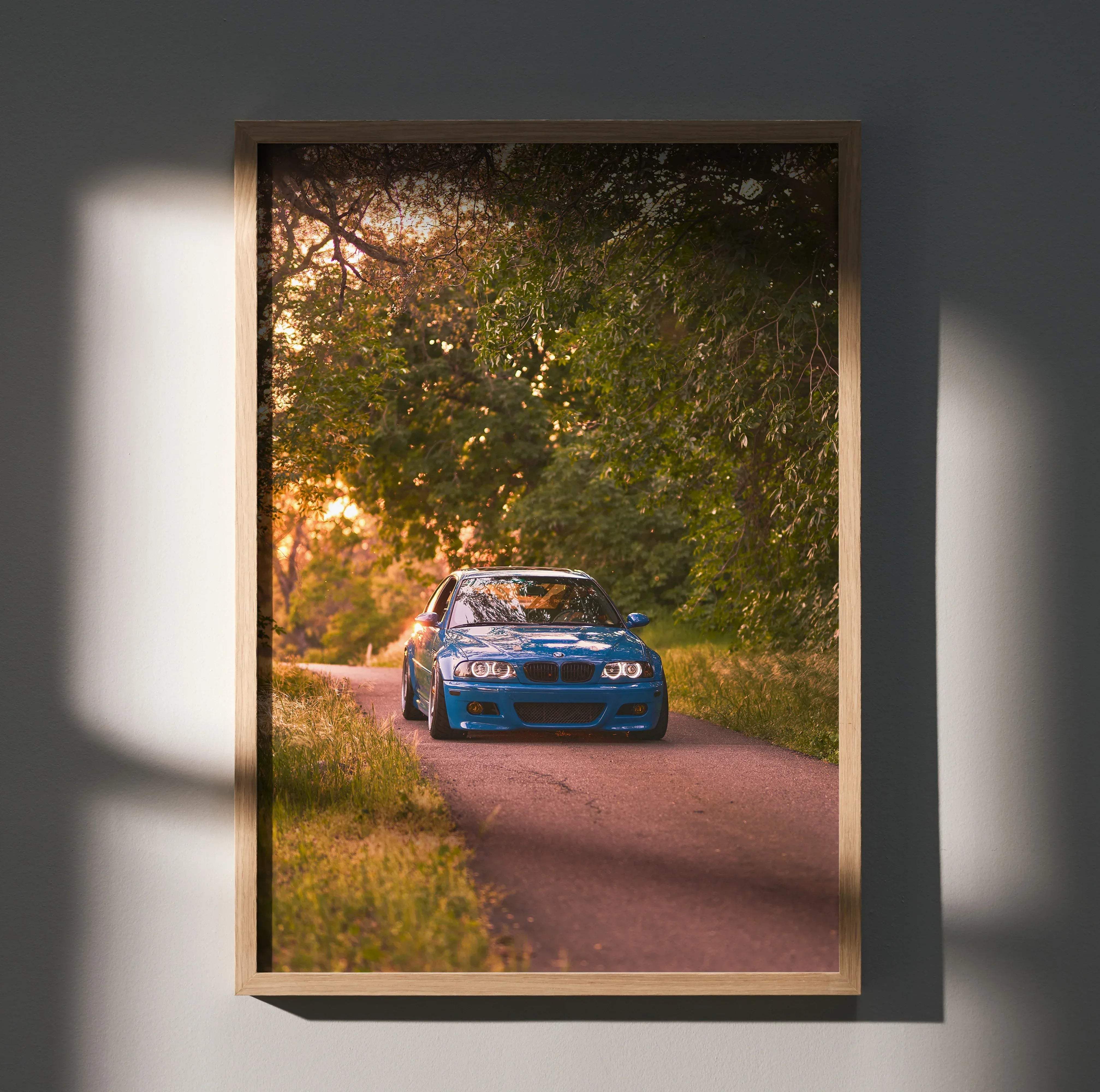 A framed BMW inspired E46 M3 art print featuring a vibrant blue car on a tree-lined road at sunset