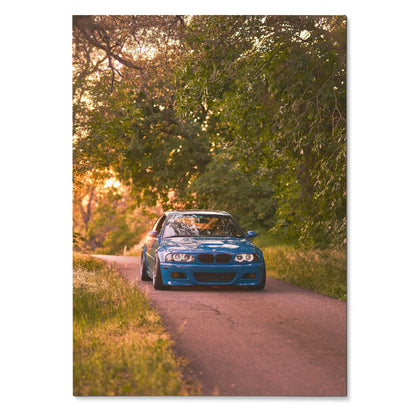 Blue BMW E46 M3 driving on tree-lined road poster, automotive wall art with warm lighting.