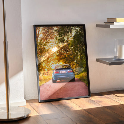 BMW E46 M3 driving on sunset-lit forest road poster wall art in black frame.