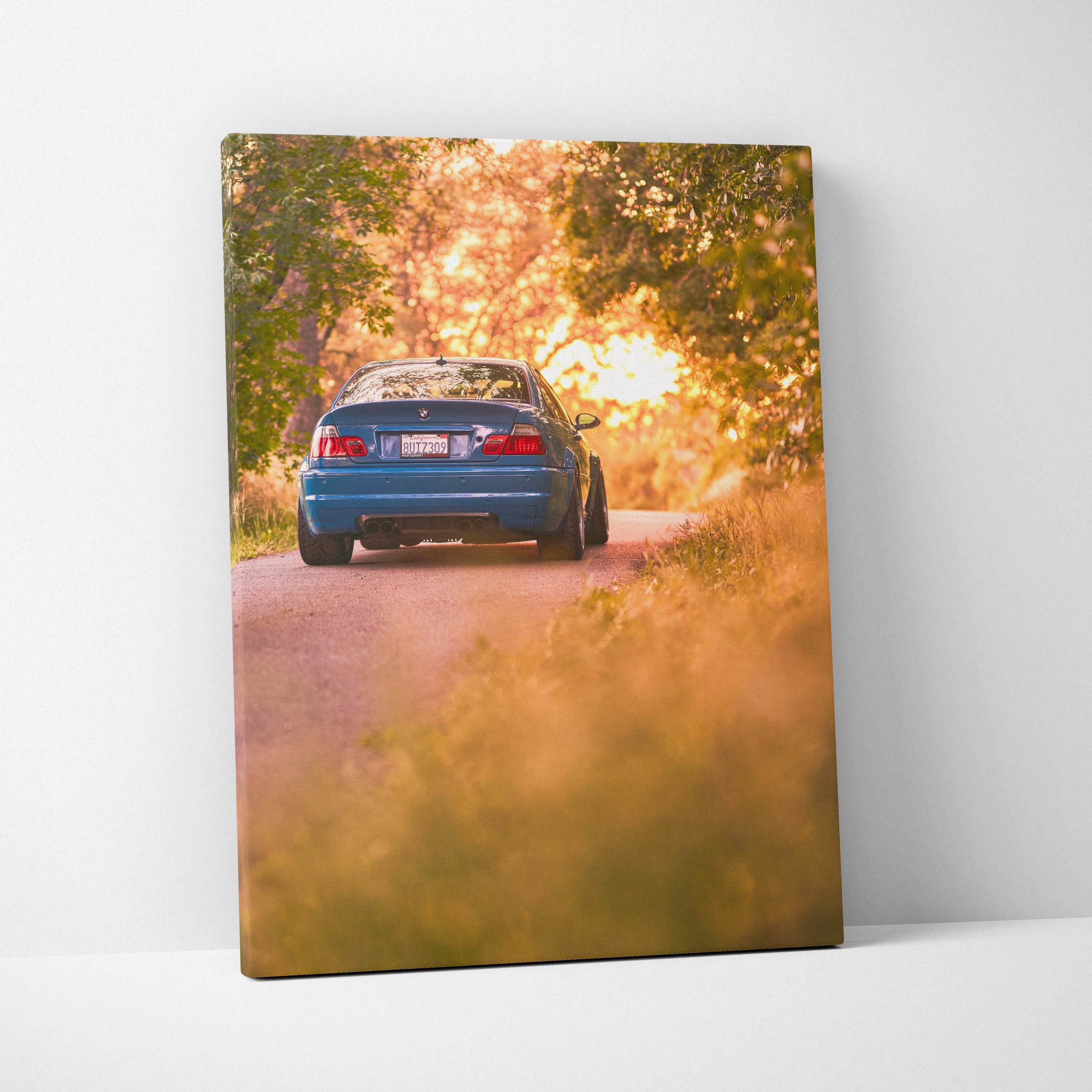 Vertical canvas wall art featuring a blue BMW E46 M3 driving on a sunlit tree-lined road at sunset.