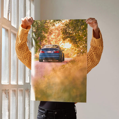 BMW E46 M3 art poster showing rear view of blue car driving on sunset-lit tree-lined road, automotive wall art.
