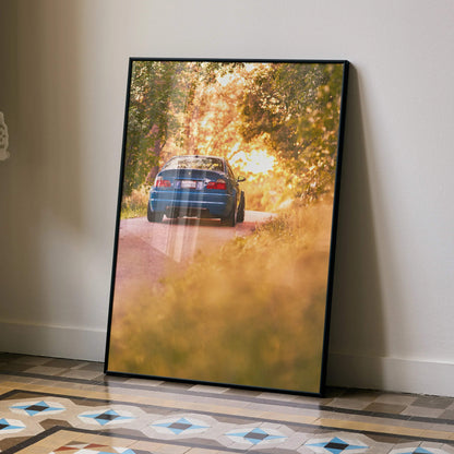 BMW E46 M3 driving on tree-lined road at sunset poster wall art with warm golden tones.