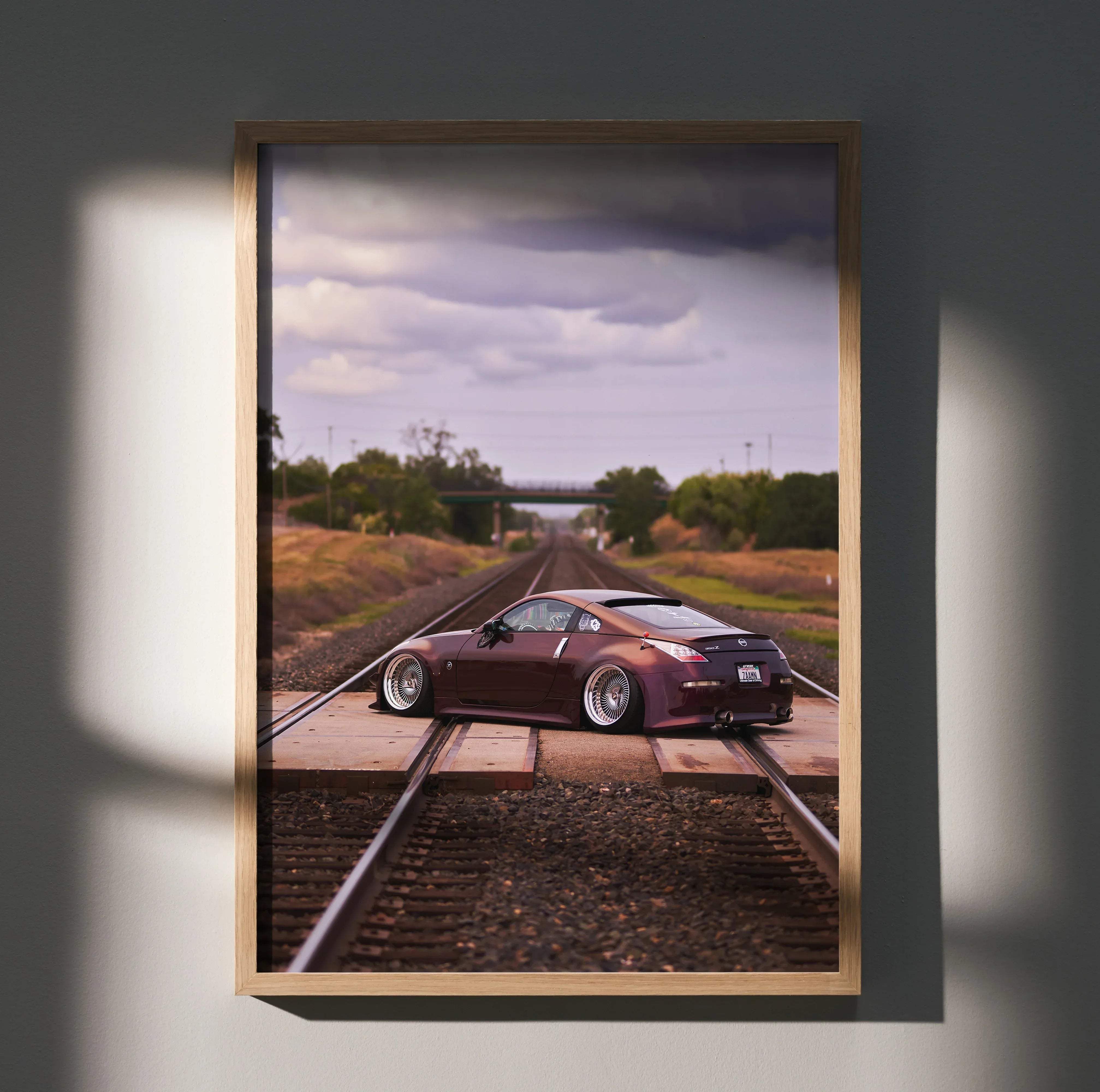 A framed photograph poster of a maroon Nissan 350Z sports car on railroad tracks under a cloudy sky for wall decor