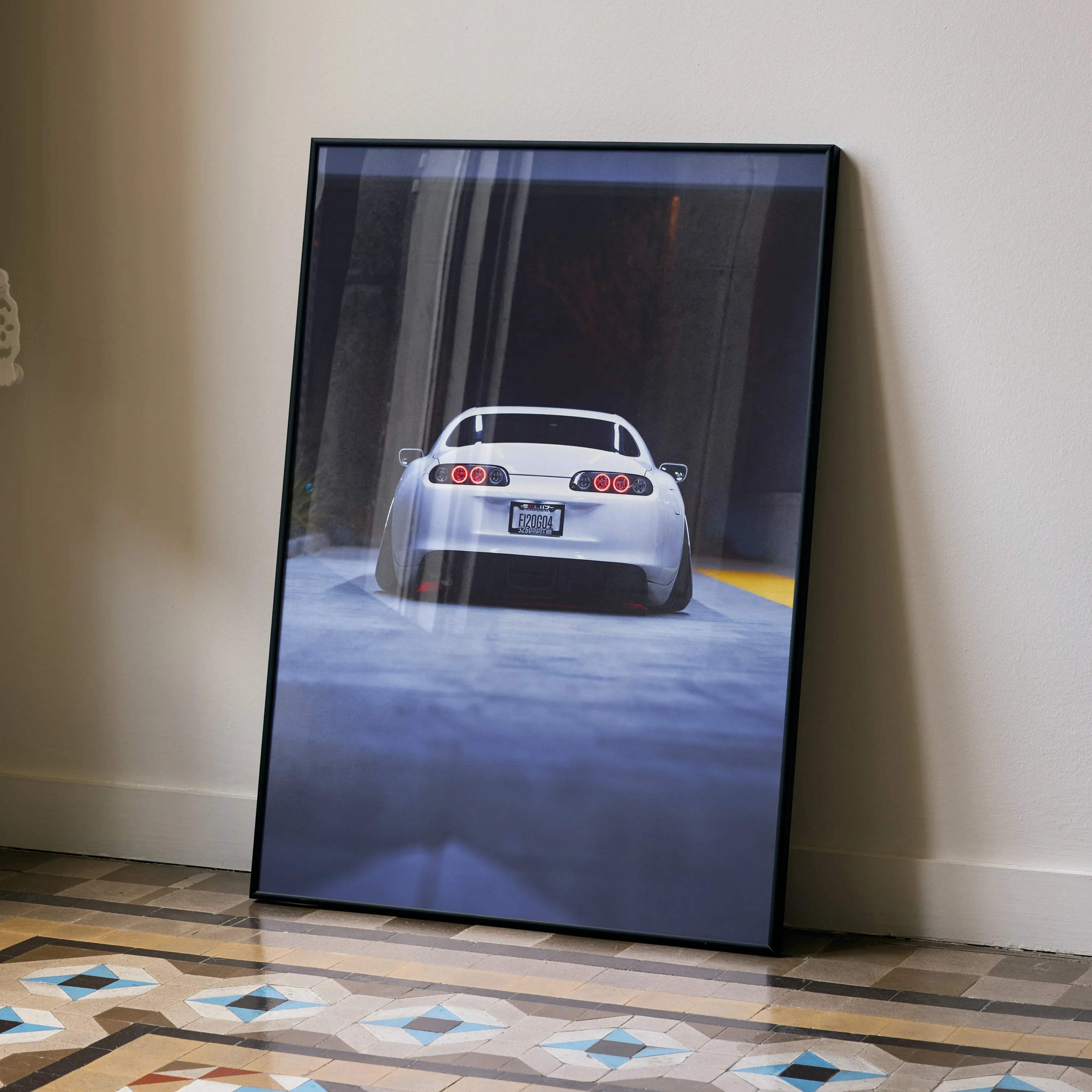 A framed art print poster of a white toyota supra mk4 with red circular taillights leaning against a wall