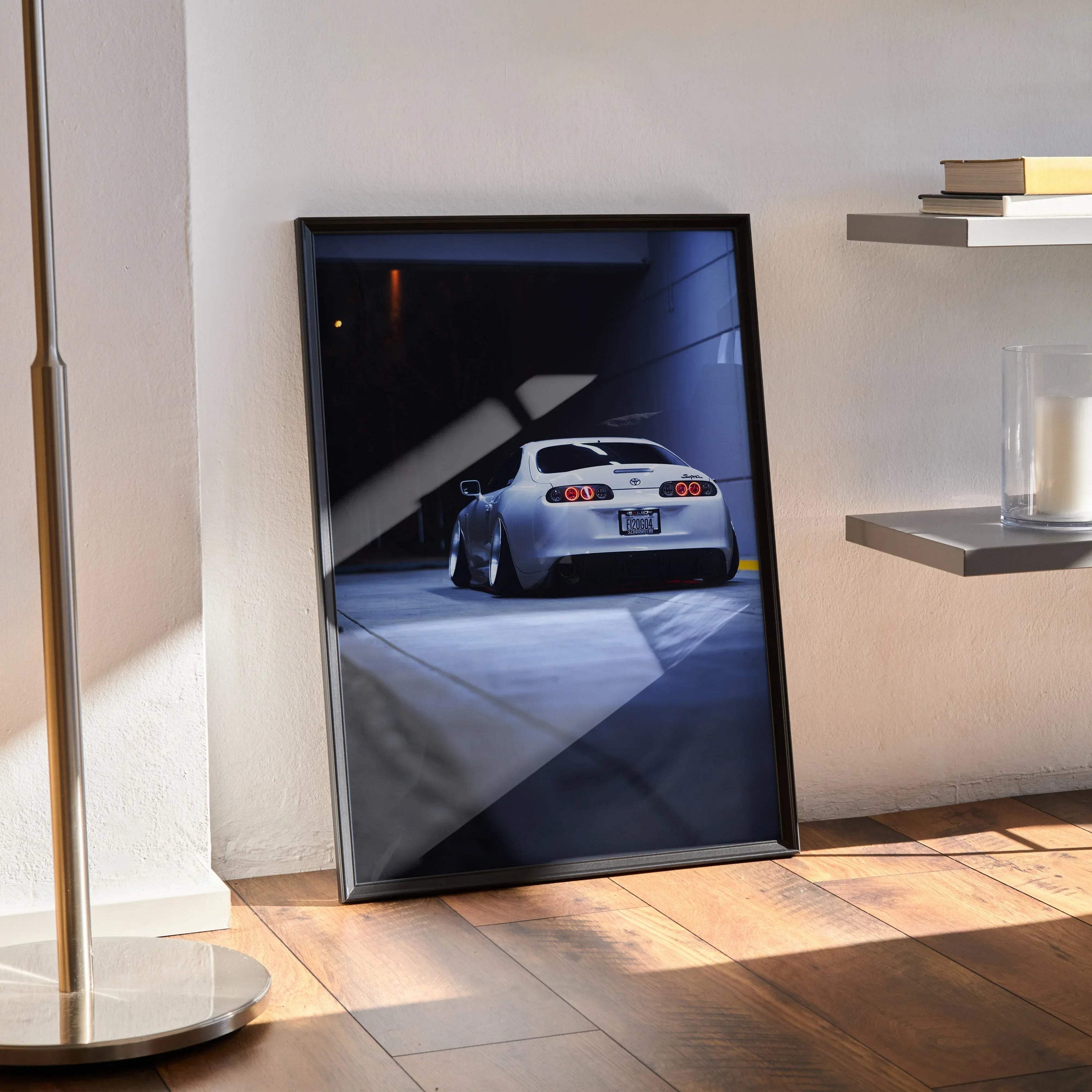 Toyota Supra MK4 rear view poster in black frame leaning on floor, automotive wall art with dramatic lighting.