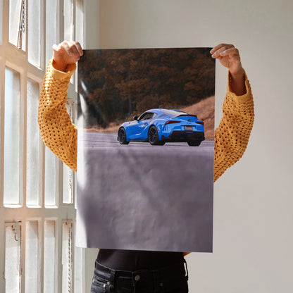 Blue Toyota Supra MK5 driving on scenic road poster wall art held against bright window light.