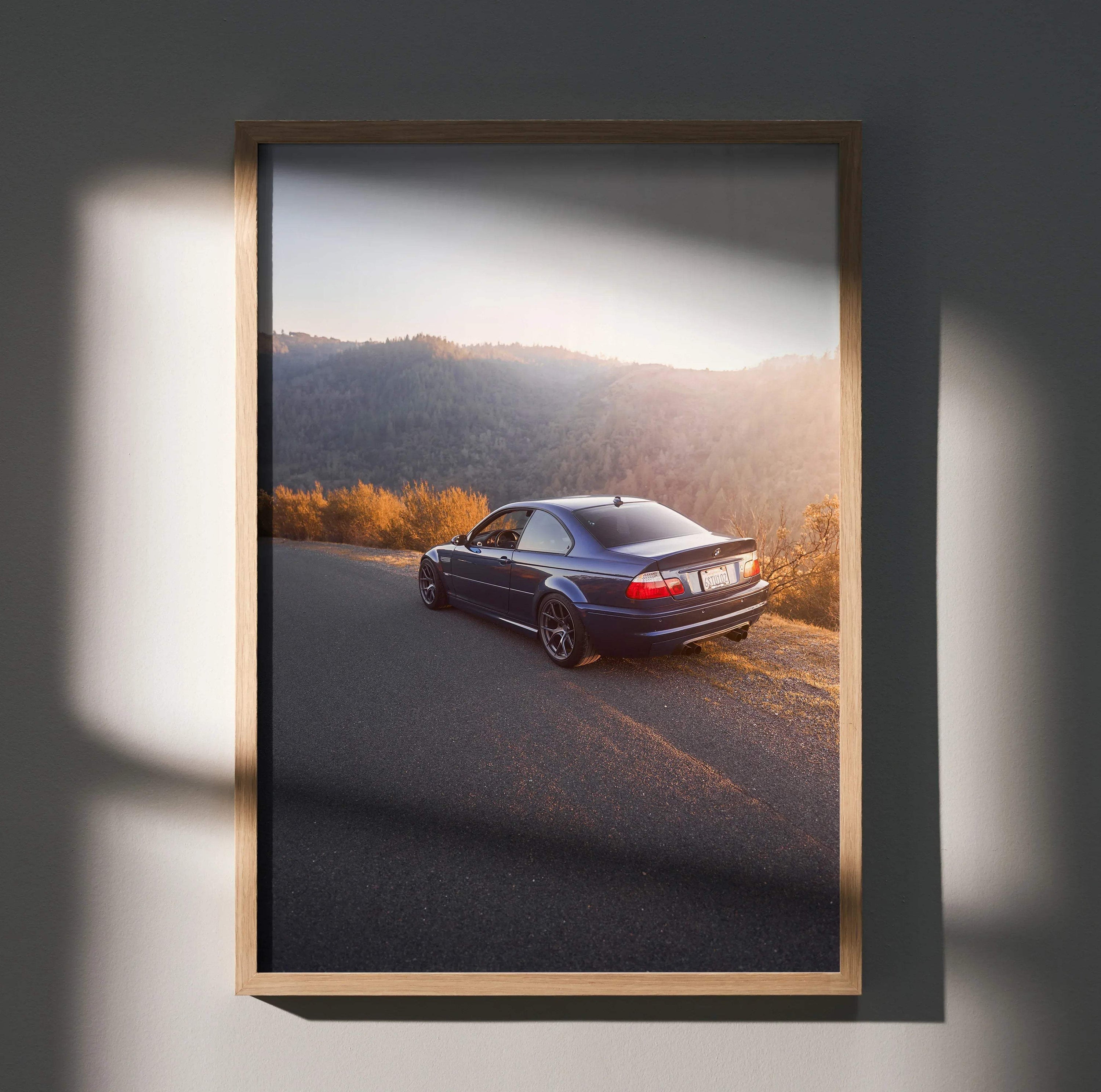BMW E46 M3 driving on scenic road at sunset poster wall art in wooden frame.
