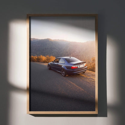 BMW E46 M3 driving on scenic road at sunset poster wall art in wooden frame.