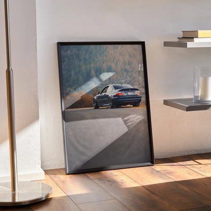 BMW E46 M3 Poster wall art with rear view of blue BMW on winding road, framed and leaning on floor.