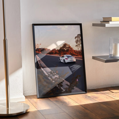 Porsche GT4 racing on a sunset track poster wall art in a black frame leaning against white wall.